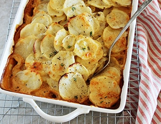 Un platillo con papas gratinadas y una cuchara sobre una rejilla y una toalla a rayas.