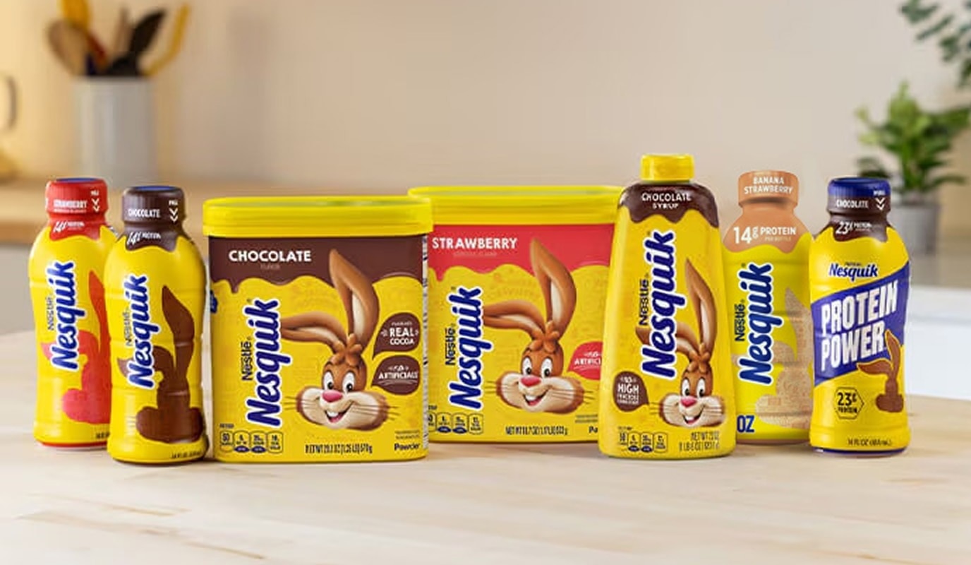 Close up of Nesquik products: bottles, powders, syrups on a beige kitchen counter with cooking utensils in the background.