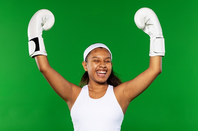 Una adolescente con camiseta sin mangas, cinta en la cabeza y guantes de boxeo, todo en blanco, levanta los brazos y celebra.