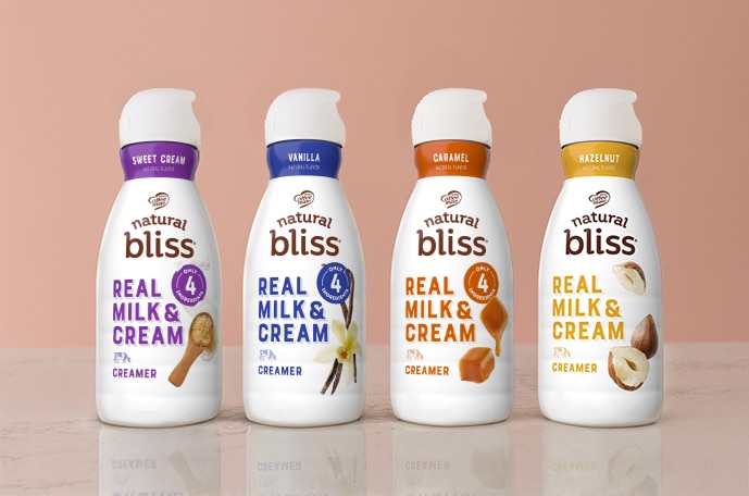 Three bottles of natural bliss plant based creamers lined up on a counter in front of a blue wall.