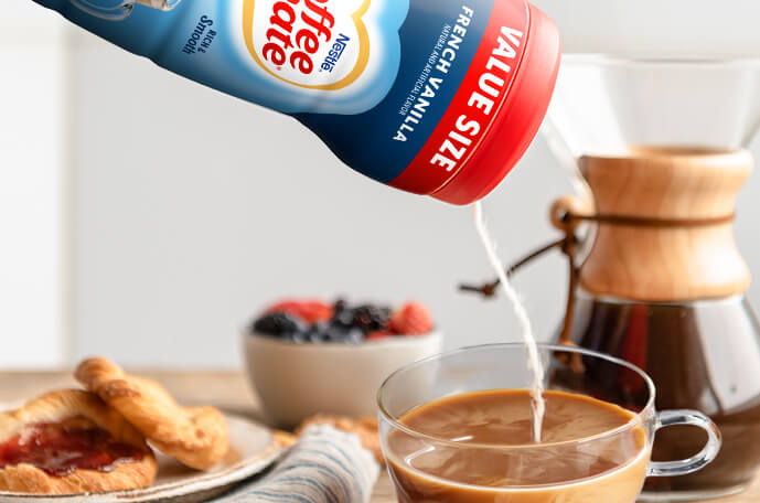 Powder pours from a canister into a mug of coffee by a pour over coffee maker, plate of pastries and bowl of fruit.