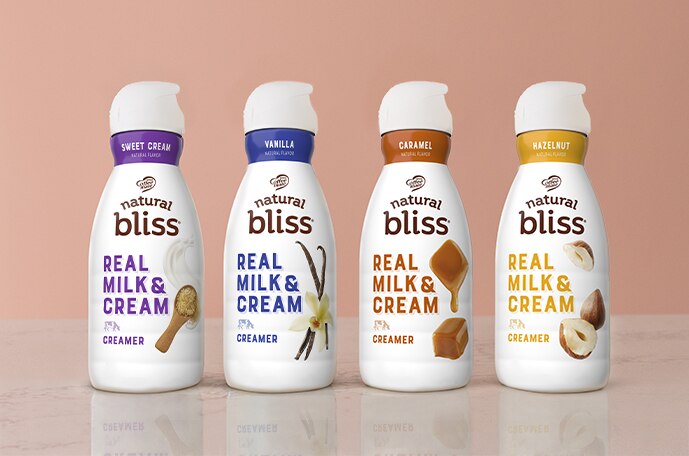 Cuatro frascos de creamers natural bliss de leche y crema auténticas alineados en una encimera frente a una pared marrón.