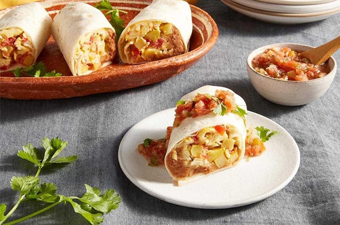 Burritos de desayuno junto a un tazón de salsa, platos y cilantro sobre una superficie gris texturizada.