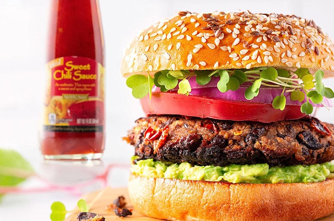 Hamburguesa sobre una tabla de cortar con brotes y salsa de chile sobre una superficie blanca.