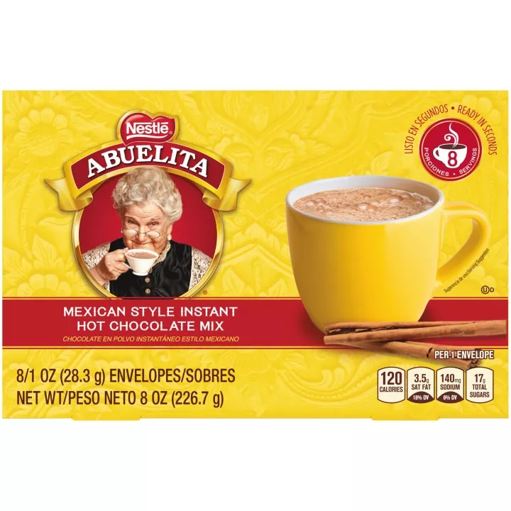 A yellow box with a red label below a yellow mug of Mexican hot chocolate behind cinnamon sticks.