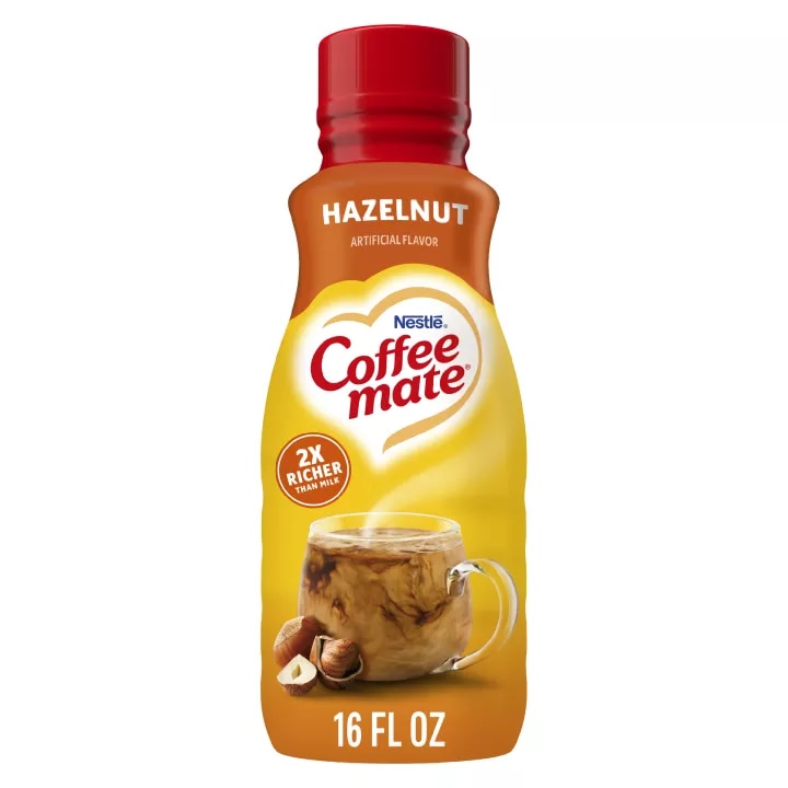 Una botella de creamer Hazelnut con una etiqueta roja encima de una taza de café, avellanas enteras y el logotipo del producto.