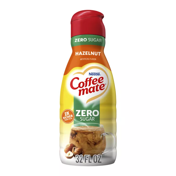 Una botella amarilla de Creamer Zero Sugar Hazelnut con carteles verdes y rojos sobre una taza de café con avellanas enteras.