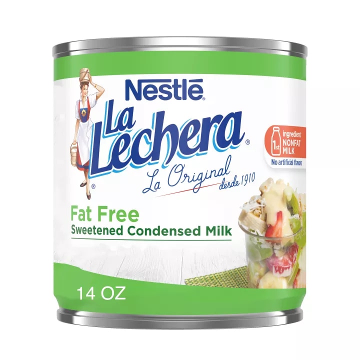 Una lata color blanco con las etiquetas color verde y el logo de La Lechera, junto a una copa de fruta rociada con leche condensada azucarada.