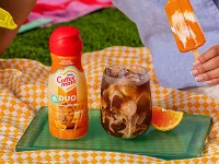Coffee Mate Orange Cream Pop Liquid Creamer on orange gingham picnic blanket with an iced coffee and orange slice next to it.