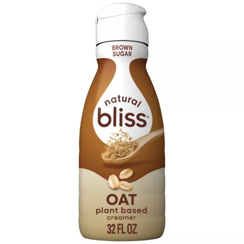 Una botella de creamer Oat Brown Sugar de natural bliss con una etiqueta marrón encima de una taza medidora con azúcar moreno.