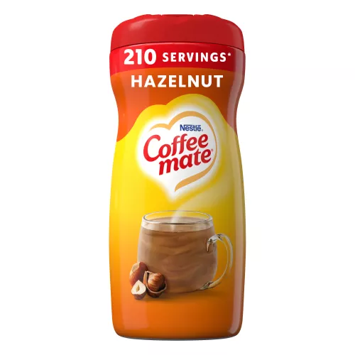 Una lata amarilla de creamer en polvo Coffee mate Hazelnut con una etiqueta roja, una taza de café, avellanas enteras y el logotipo del producto.
