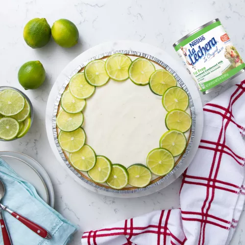 Una lata color blanco con las etiquetas color verde y el logo de La Lechera, junto a una copa de fruta rociada con leche condensada azucarada.
