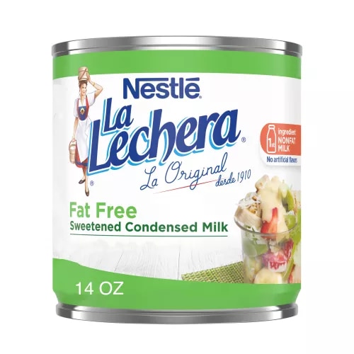 Una lata color blanco con las etiquetas color verde y el logo de La Lechera, junto a una copa de fruta rociada con leche condensada azucarada.