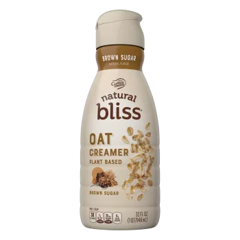 Una botella de creamer Oat Brown Sugar de natural bliss con una etiqueta marrón encima de una taza medidora con azúcar moreno.