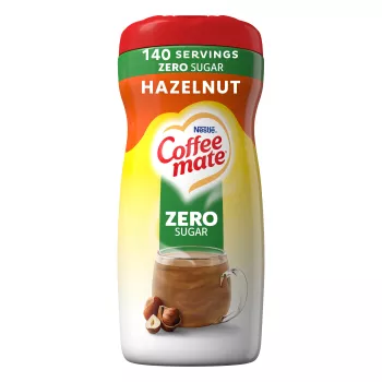 Una lata de creamer en polvo Hazelnut con una etiqueta verde que dice "sin azúcar" encima de una taza de café, avellanas enteras y el logotipo del producto.