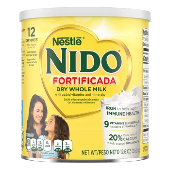Una lata de Nido Fortificada con una etiqueta amarilla, una tapa blanca y un corazón dorado sobre un vaso de leche vertida.