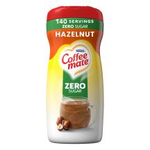 Una lata de creamer en polvo Hazelnut con una etiqueta verde que dice "sin azúcar" encima de una taza de café, avellanas enteras y el logotipo del producto.