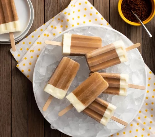Cinco paletas heladas bicolor (café y crema) sobre hielo en plato, servilleta con lunares