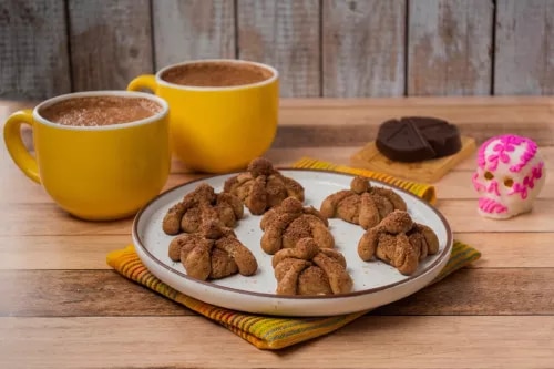 Photo: two yellow mugs of cocoa, plate of chocolate crescent cookies, pink donut on wood.