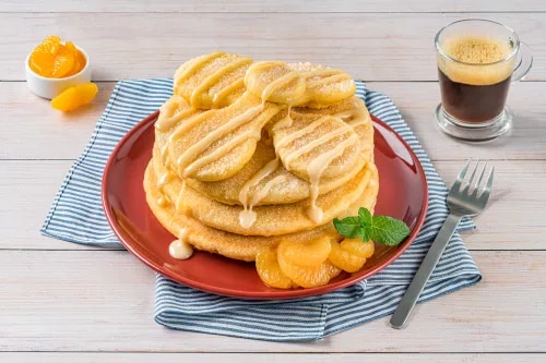 Photo of stacked pancakes with syrup, peach slices, mint on red plate, espresso