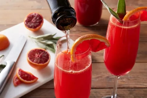 Two blood-orange cocktails in flutes, one being poured, garnished with orange slice and rosemary.