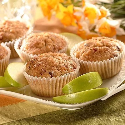 Fotografía de cuatro magdalenas con azúcar en plato, rodajas de manzana verde y flores