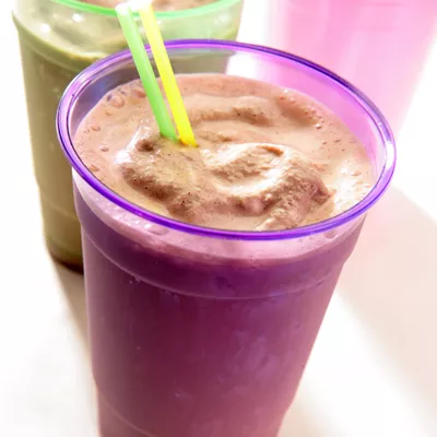 Foto de batido cremoso rosa en vaso morado con dos pajillas, batidos verde y rosa al fondo