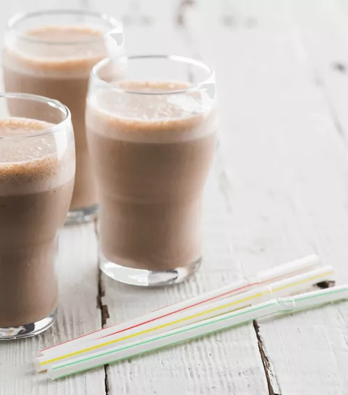 Fotografía: tres vasos con batido de chocolate espumoso sobre mesa blanca y pajillas pastel.
