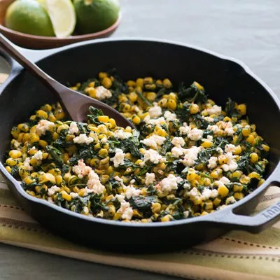 Foto: sartén con espinacas, maíz y queso desmenuzado; cuchara de madera y limas