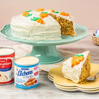 Cream-frosted carrot cake on green stand; slice on yellow plate, frosting cans and pastel eggs